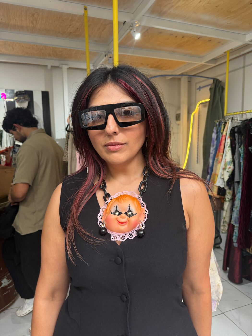 Karishma in a black vest, red-streaked hair, oversized black shades and a necklace with a vintage doll face as the pendant, standing in a vintage clothing shop.
