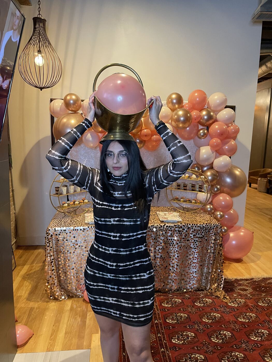 Karishma in a black and white striped dress and clear glasses, balancing a brass pot holding a pink balloon on her head in front of a balloon arch.
