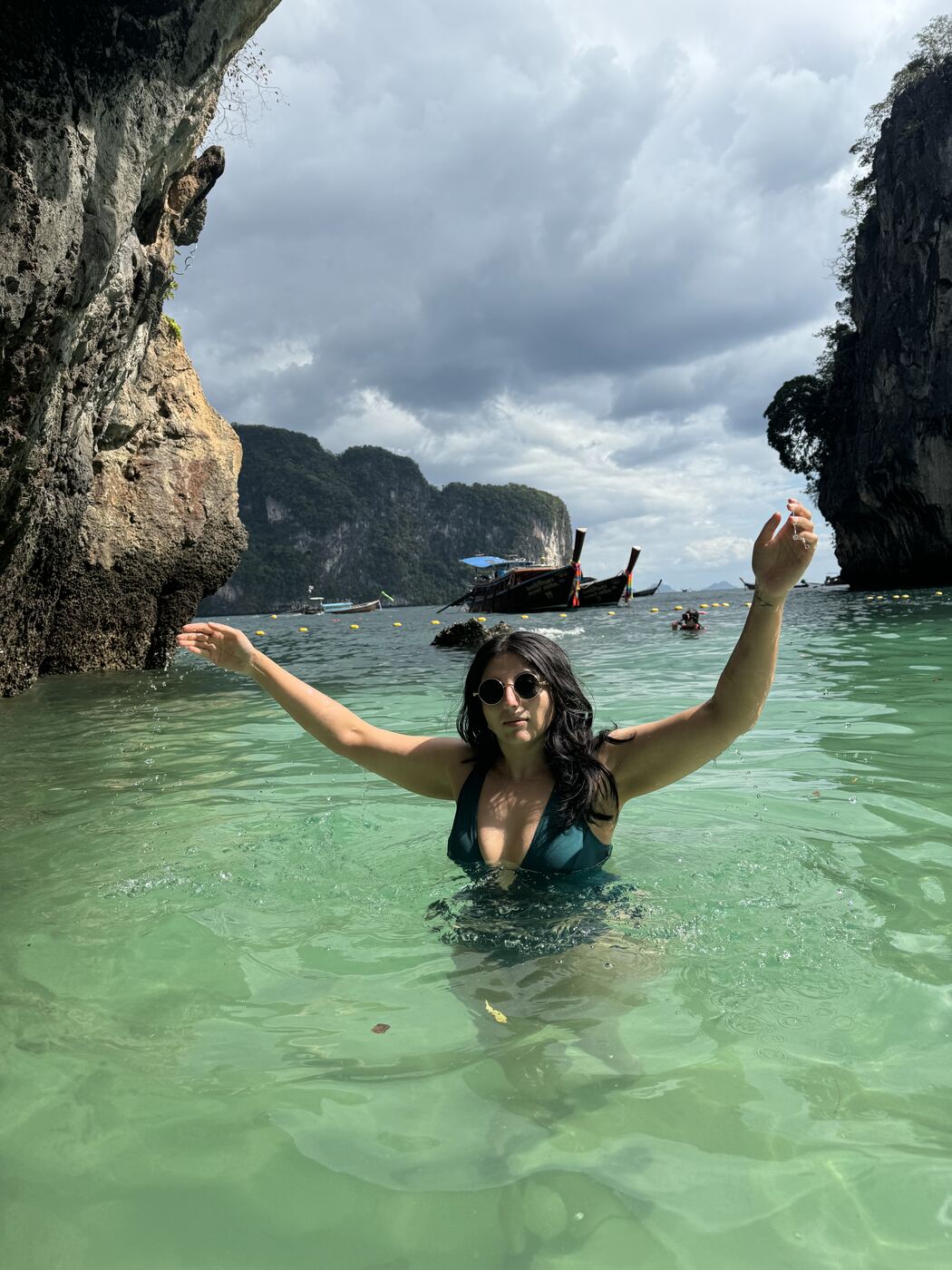 Karishma waist deep in clear green water with arms raised, limestone cliffs and longtail boats behind her.