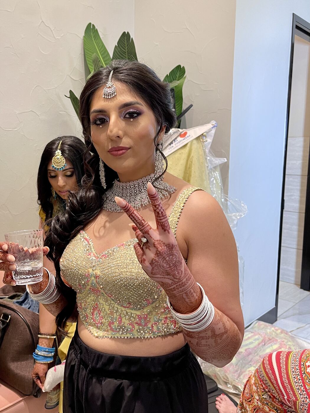 Karishma dressed up in an embellished gold blouse, a statement choker and a maang tikka, with henna on her hands, holding up a peace sign.